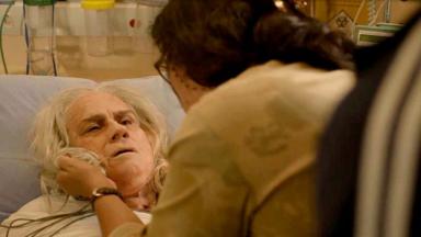 Lurdes, de costas, segura Kátia pelo colarinho enquanto ela está deitada num leito do hospital, de frente 