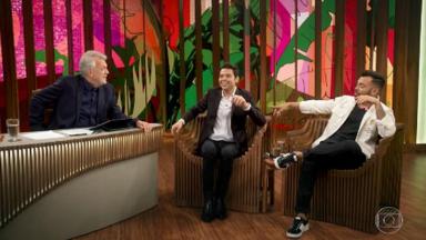 Pedro Bial sentado na bancada do "Conversa com Bial", ao lado de Gustavo Mendes e Thiago Brava, também sentados 