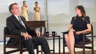 Jair Bolsonaro sentado numa cadeira, ao lado de Luciana Gimenez, também sentada 