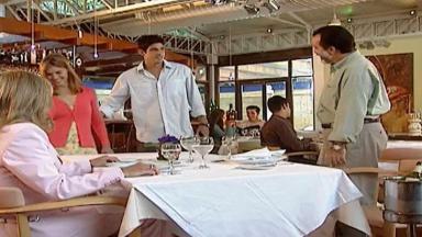 Cena de Laços de Família com Helena sentada à mesa, Miguel, Edu e Camila de pé, no restaurante 