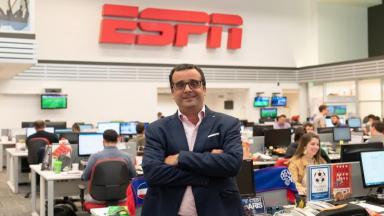 Carlos Maluf na redação da ESPN Brasil, de pé e com os braços cruzados. Na redação, há jornalistas sentados em mesas com computadores. 