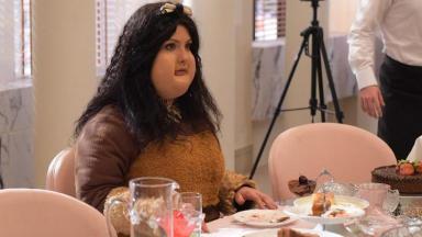 Camila Rodrigues caracterizada como Sophia muito gorda e comendo sentada numa mesa 