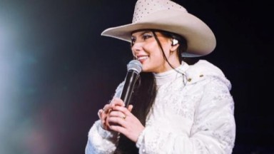 Ana Castela de rabo de cavalo, com casaco e chapeu brancos, segurando microfone e olhando para o lado 