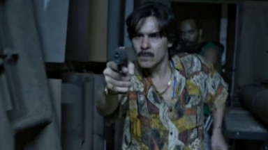 André Gonçalves apontando arma, vestindo camisa estampada, em cena de Impuros 