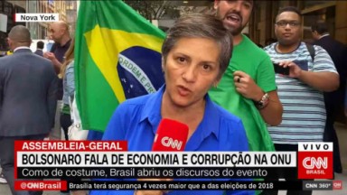 Repórter segurando microfone diante das câmeras com a presença de homem segurando bandeira do Brasil e gritando atrás dela 