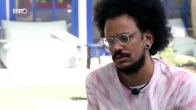 João Luiz está com semblante reflexivo de óculos e blusa branca com detalhes em rosa na área externa da casa do BBB21 