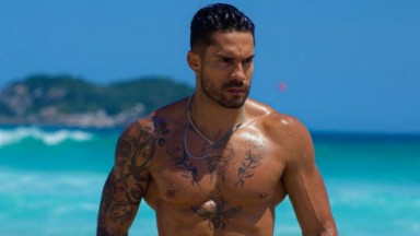 Bil Araújo sem camisa, com expressão séria, em praia 