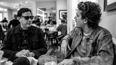 Foto em preto e branco de Bob Saget e John Mayer sentados lado a lado, conversando 