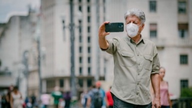 Caco Barcellos se filmando nas ruas com o celular, vestindo camisa cinza 