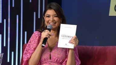 Amanda Françozo sorrindo com seu primeiro livro em mãos no palco da TV Aparecida 