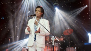 Roberto Carlos de roupa branca no palco, cantando perto de microfone 