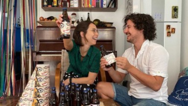 Daphne Bozaski e Gustavo Araujo sorrindo um para o outro e colando adesivos em garrafas de vidro 