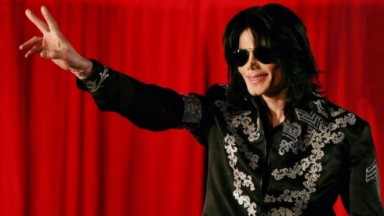 Michael Jackson de roupa preta e óculos escuros, sorrindo e fazendo sinal de jóia 