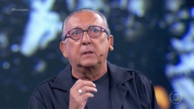 Galvão Bueno falando, usando óculos e roupas pretas 
