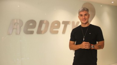 Ivan Moré de roupa preta e sorriso tímido, posando perto de símbolo da RedeTV! 