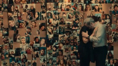 Cena de casal se abraçando perto de quadro com fotos de vítimas em Todo Dia a Mesma Noite 