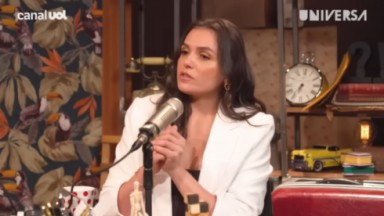 Monica Iozzi falando em microfone, de blusa preta e blazer branco 