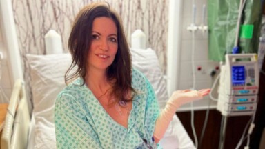 Deborah James sentada e sorrindo em cama de hospital, com a mão esquerda levantada 