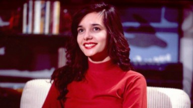 Daniella Perez de blusa de frio e batom vermelhos e cabelos soltos, sentada e sorrindo 