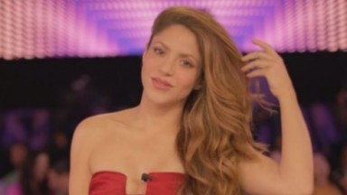  Shakira de cropped tomara que caia vermelho e cabelos soltos, passando a mão nas madeixas 