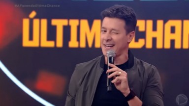 Rodrigo Faro de blusa preta e casaco cinza, piscando e segurando microfone 