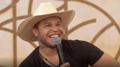 Tierry de chapéu bege e camiseta preta, sorrindo e segurando microfone 