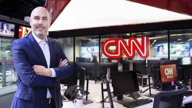 Douglas Tavolaro com os braços cruzados posando para fotos tendo ao fundo a redação da CNN Brasil 