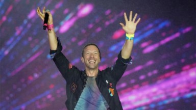 Vocalista do Coldplay sorrindo com as mãos para o alto 
