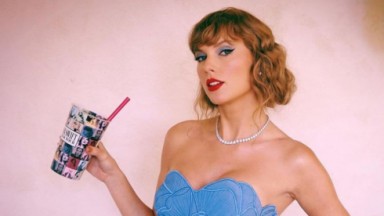 Taylor Swift de roupa tomara-que-caia azul, posando sem sorrir e segurando copo da The Eras Tour 