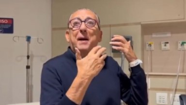 Galvão Bueno de roupa azul marinho e óculos, falando e gesticulando em hospital 