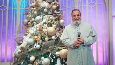 Padre Antonio Maria posado em frente a uma árvore de Natal, segurando um microfone 