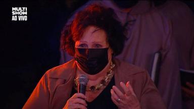 Déa Lúcia emocionada, com máscara no rosto e microfone na mão 