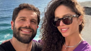 Duda Nagle e Michele Balsamão Morais sorrindo para selfie perto do mar, ele de preto e ela de roxo e óculos escuros 