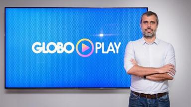 Érick Bretas de braços cruzados e de pé posa para foto com um telão ao fundo com o logo do Globoplay 