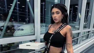 Fernanda Medrado em aeroporto, com caixa na mão e roupa preta, fazendo carão, em pé e com fones de ouvido 