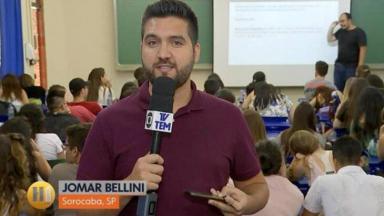 Jomar Bellini em uma sala de aula cheia de estudante e professor, durante link, segurando microfone,  