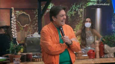 Faustão de blusa verde e jaqueta laranja rindo e segurando microfone 