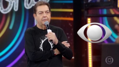 Faustão de braços cruzados, roupa preta e microfone na mão no cenário do programa na Globo 