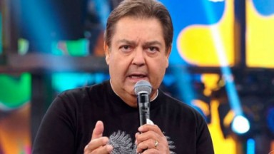 Faustão de jaqueta preta, segurando microfone, no cenário do Domingão, da Globo 