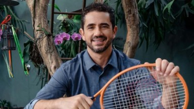Felipe Andreoli sorrindo, de camisa jeans, segurando raquete de tênis 