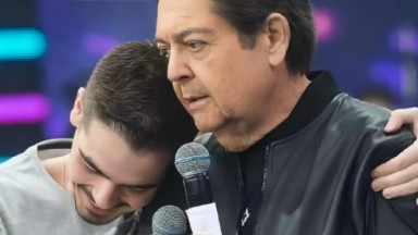 João Guilherme Silva e Faustão abraçados e sorrindo, com microfones 