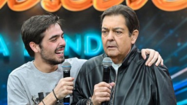 João Guilherme Silva e Faustão abraçados, segurando microfone, o filho rindo e o pai sério 