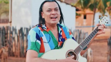 Frank Aguiar sorrindo e tocando violão, com camisa colorida 
