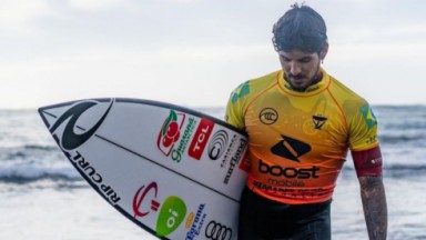 Gabriel Medina com prancha de surfe, saindo do mar, de cabeça baixa 