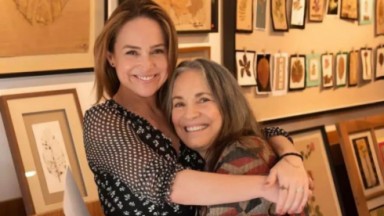 Gabriela Duarte e Regina Duarte abraçadas e sorrindo para foto perto de quadros 