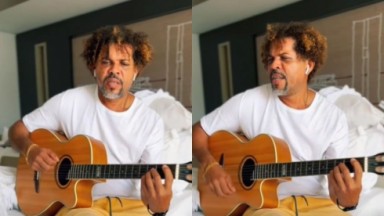 Givaldo Alves de bermuda amarela e camiseta branca sentado em cama tocando violão 