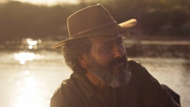 Marcos Palmeira como José Leôncio em Pantanal. Ele está sentado em seu barco na beira do rio 