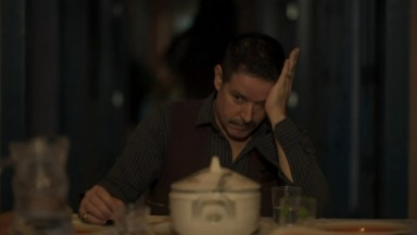 Murilo Benício em cena como Tenório na novela Pantanal. Ele está sentado e com o braço em cima da mesa da cozinha 