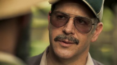Murilo Benício como Tenório em Pantanal. O personagem está em pé, usando óculos e boné 