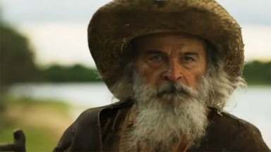 Osmar Prado em cena como Velho do Rio na novela Pantanal. Ele está em pé perto do rio 